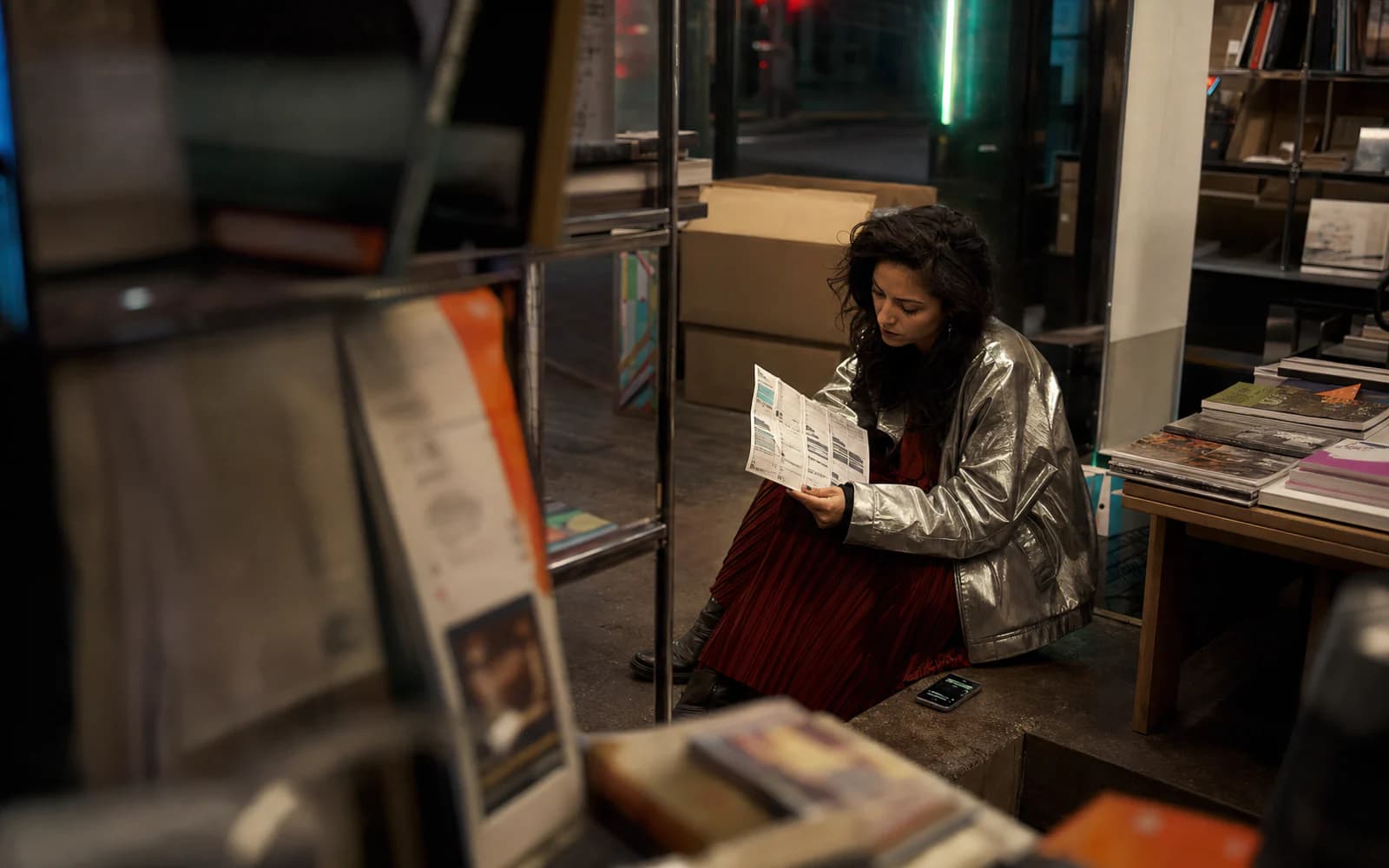 Adult woman in a silver jacket and red dress reviewing a pop-up schedule in a bookshop at night
