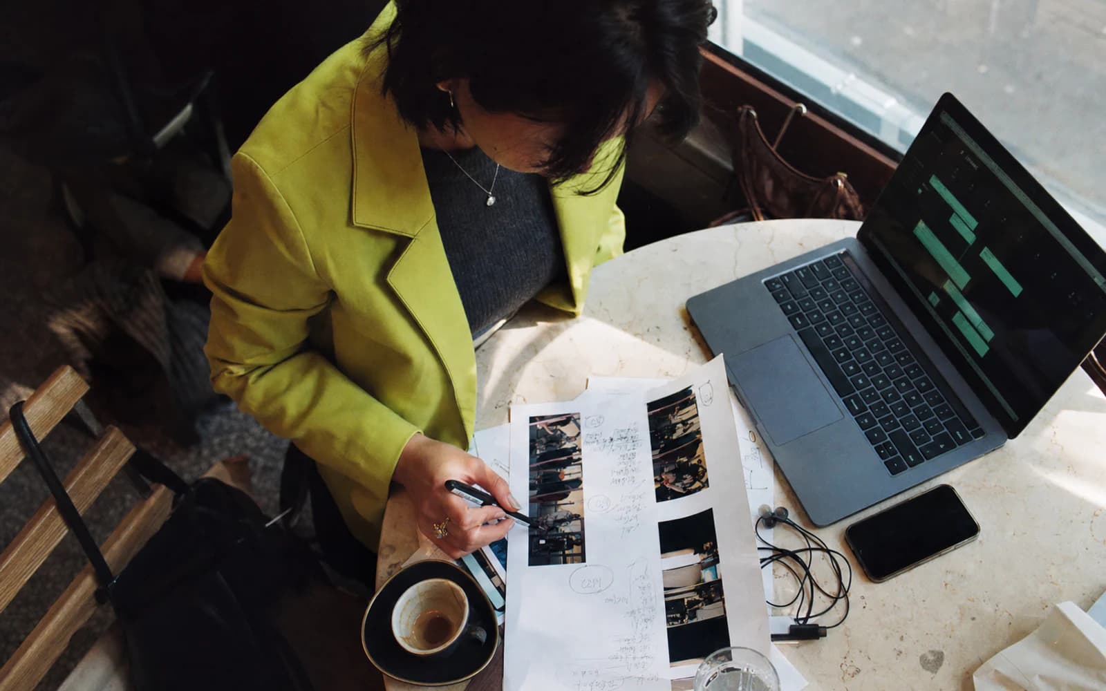 Overhead view of an adult woman in a lime blazer reviewing printed material and a laptop at a café table